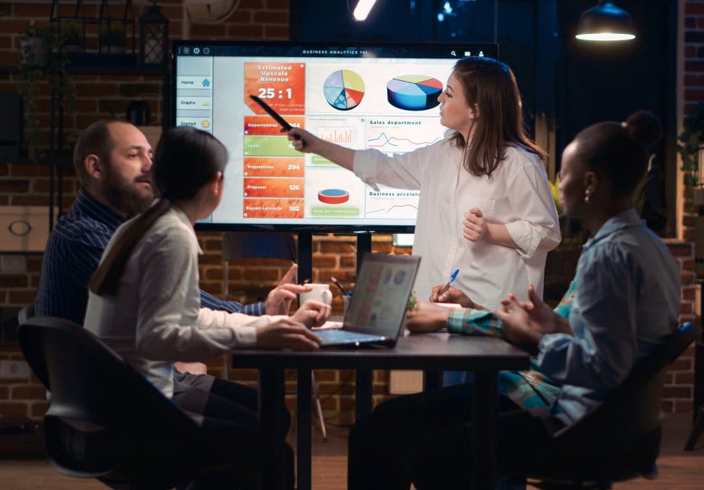 A diverse group of individuals gathered around a table with a projector screen displaying information.