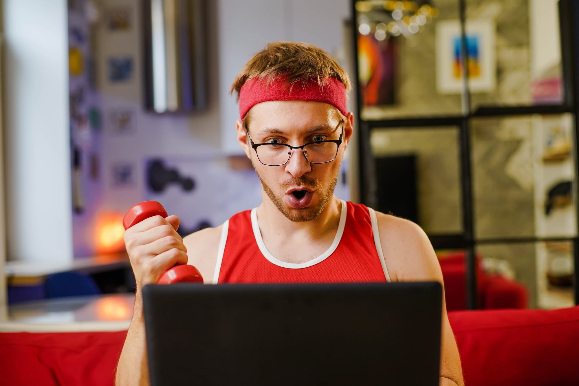 Funny young man geek with dumbbells watching training program on laptop online.